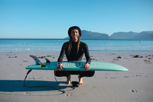 Портрет улыбающегося серфера, приседающего с доской для серфинга на пляже на фоне ясного голубого неба с копировальным пространством. хобби и водные виды спорта. — стоковое фото