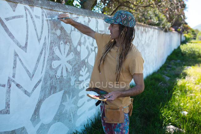 Männlicher Hipster-Künstler hält Palette, während er abstrakte Wandmalerei an die Wand malt. Street Art und Geschicklichkeit. — Stockfoto