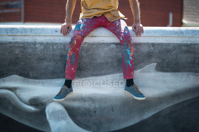 Low section of male artist with messy pant sitting on wall with creative mural painting of wall. street art, creativity and skill. — Stock Photo