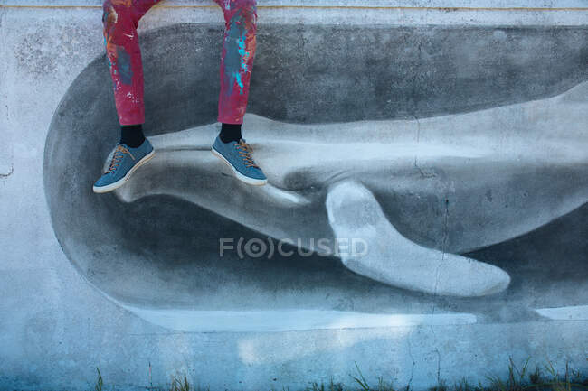 Sección baja de artista masculino con pantalón desordenado sentado en la pared con hermosa pintura mural de la pared. arte urbano, creatividad y habilidad. - foto de stock
