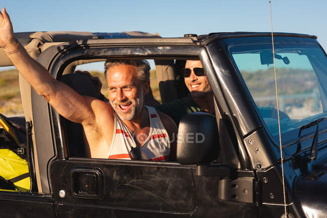 Felice coppia maschile gay caucasica che saluta, seduta in auto nella giornata di sole al mare. estate viaggio su strada e vacanza nella natura. — Foto stock