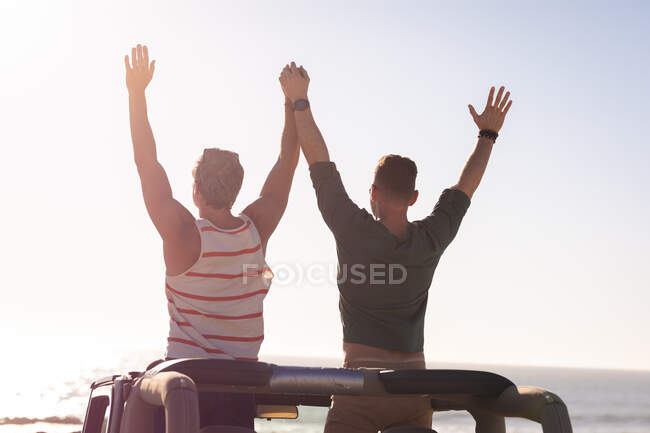 Rear view of caucasian gay male couple raising arms and holding hands, sitting on car in sun by sea. summer road trip and holiday in nature. — Stock Photo