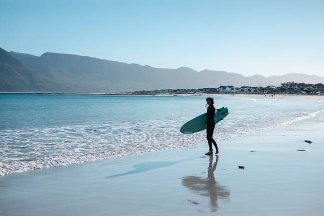 Hombre llevando tabla de surf en la orilla en la playa contra el cielo azul con espacio de copia. pasatiempos y deportes acuáticos. - foto de stock