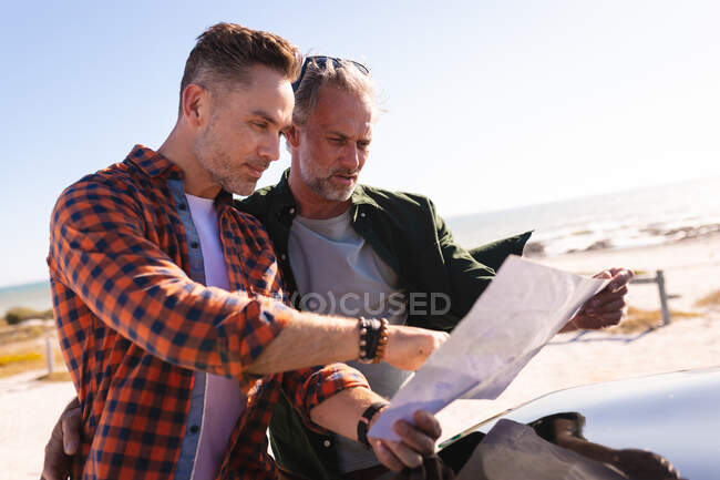 Feliz pareja gay caucásica leyendo mapa en un coche en la playa. viaje por carretera de verano y vacaciones en la naturaleza. - foto de stock