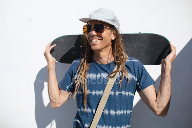 Smiling fashionable hipster man wearing sunglasses and cap with skateboard behind head against wall. lifestyle, fashion and sport. — Stock Photo