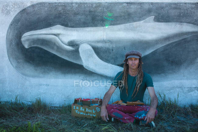 Fier artiste hipster masculin assis jambes croisées devant sa peinture murale de baleine sur le mur. street art et savoir-faire. — Photo de stock