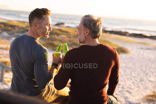 Happy caucasian gay male couple drinking bottles of beer and talking at seaside. summer road trip and holiday in nature. — Stock Photo