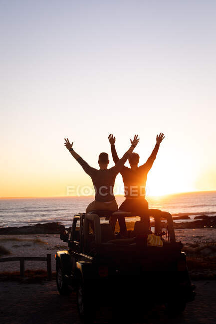 Vue arrière du couple gay caucasien assis sur le toit de la voiture levant les bras au coucher du soleil au bord de la mer. road trip d'été et vacances dans la nature. — Photo de stock