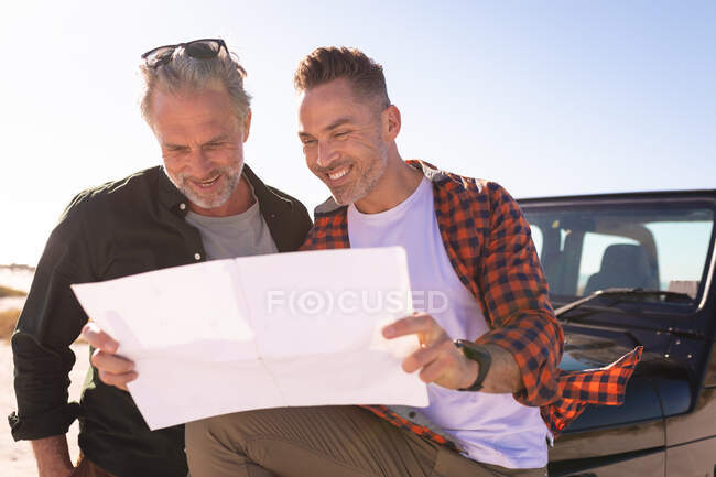 Feliz caucasiano gay casal leitura mapa, sorrindo por um carro à beira-mar. viagem de verão e férias na natureza. — Fotografia de Stock