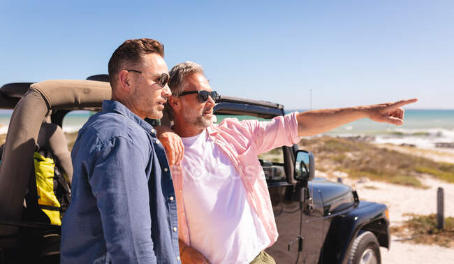 Happy caucasian gay male couple embracing and pointing by a car at seaside. summer road trip and holiday in nature. — Stock Photo