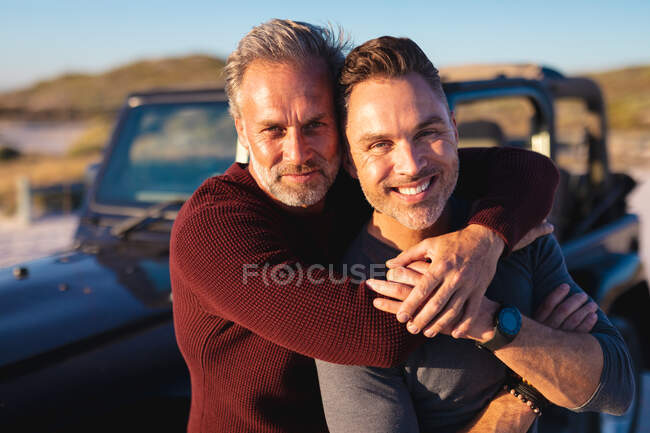 Retrato de casal masculino gay caucasiano sorridente abraçando por um carro no dia ensolarado à beira-mar. viagem de verão e férias na natureza. — Fotografia de Stock