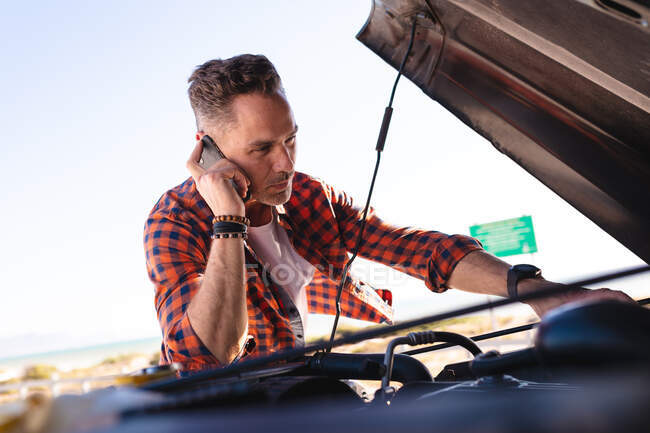 Homem caucasiano estressado falando no smartphone, por carro quebrado com tampa aberta à beira-mar. viagem de verão e férias na natureza. — Fotografia de Stock