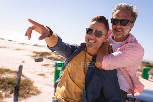 Sonriente pareja gay caucásica abrazando y señalando, disfrutando de la vista en la playa. viaje por carretera de verano y vacaciones en la naturaleza. - foto de stock