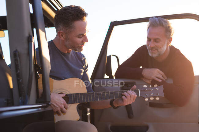 Heureux couple gay gay caucasien jouant de la guitare et souriant, traînant en voiture au bord de la mer. road trip d'été et vacances dans la nature. — Photo de stock