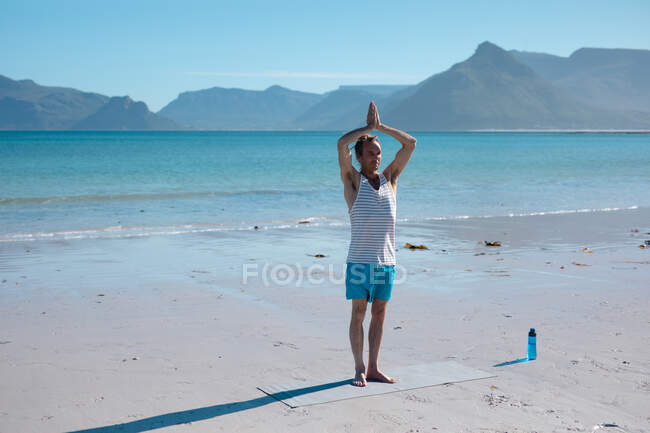 Hombre practicando yoga con las manos apretadas sobre la cabeza en la alfombra en la playa durante el día soleado. fitness y estilo de vida saludable. - foto de stock