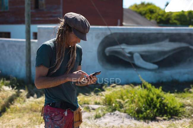 Мужчина фреска художник, использующий смартфон, стоя возле китовой живописи на стене в солнечный день. технологии, стрит-арт и мастерство. — стоковое фото