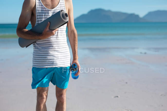 Midsection do homem que prende a esteira do ioga e a garrafa de água na praia no dia ensolarado. fitness e estilo de vida saudável. — Fotografia de Stock