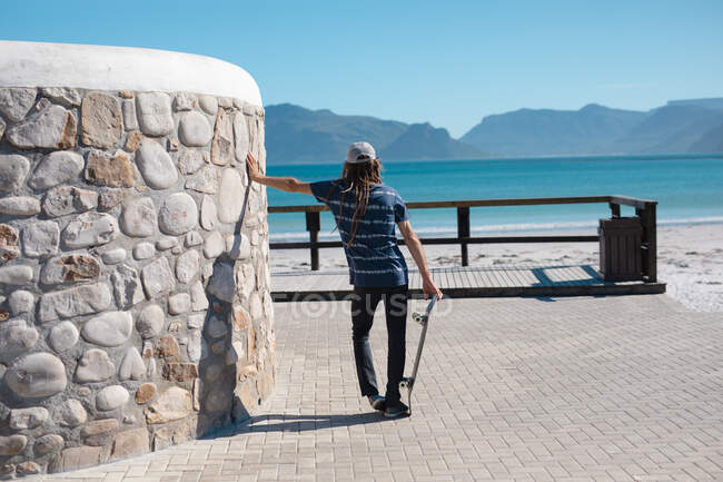 Вид сзади на человека, держащего скейтборд, опирающегося на стену на набережной, когда он смотрит на море. образ жизни и спорт. — стоковое фото