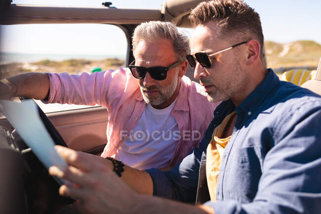Focused caucasian gay male couple reading map sitting in car at seaside. summer road trip and holiday in nature. — Stock Photo