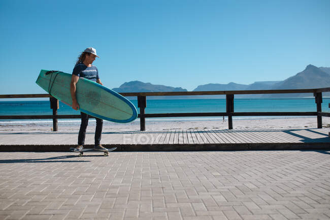 Homem carregando prancha de surf enquanto skate no calçadão pela praia contra o céu azul com espaço de cópia. hobbies, estilo de vida e esporte. — Fotografia de Stock