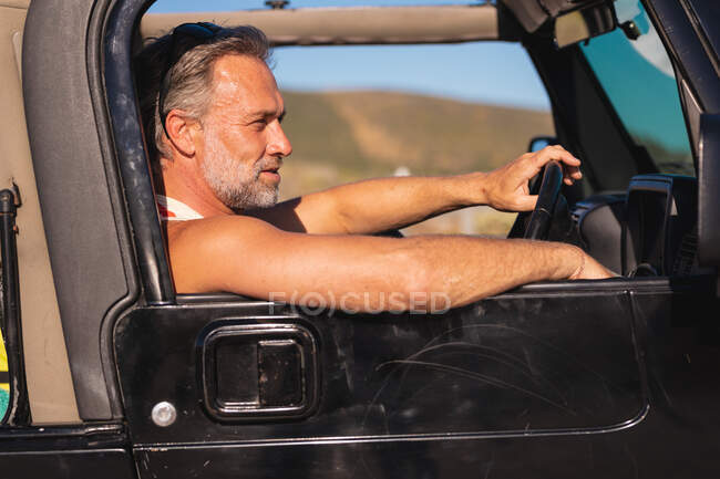 Happy caucasian man sitting in car at seaside on sunny day. summer road trip and holiday in nature. — Stock Photo