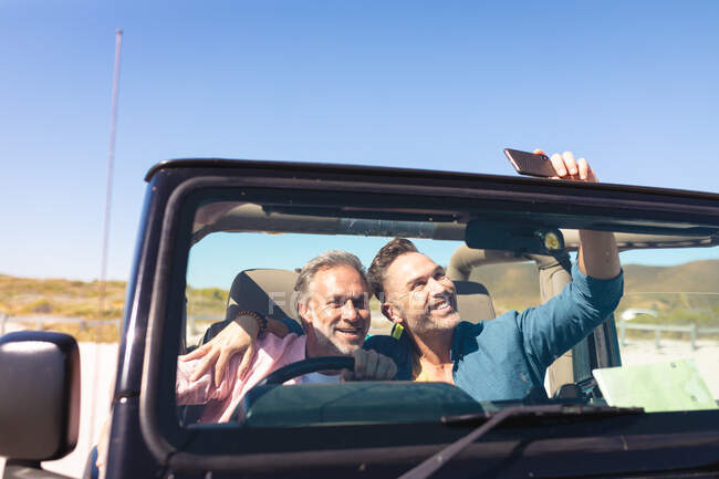 Sonriendo caucásica pareja gay macho tomando selfies sentado en el coche en la playa. viaje por carretera de verano y vacaciones en la naturaleza. - foto de stock