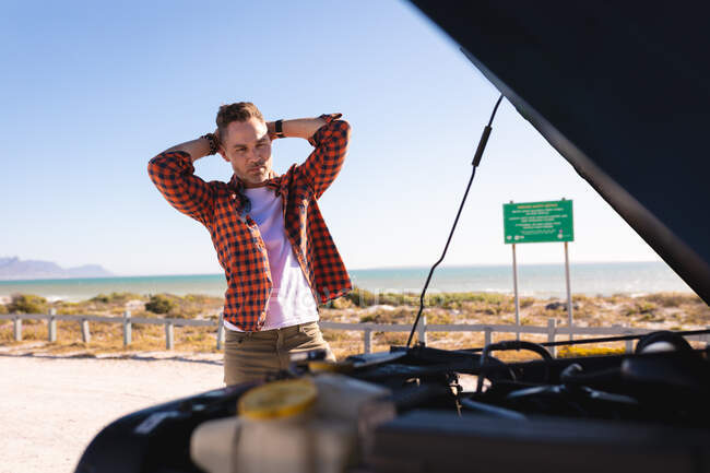 Stressed caucasian man holding his head looking at broken down car with open bonnet at seaside. summer road trip and holiday in nature. — Stock Photo