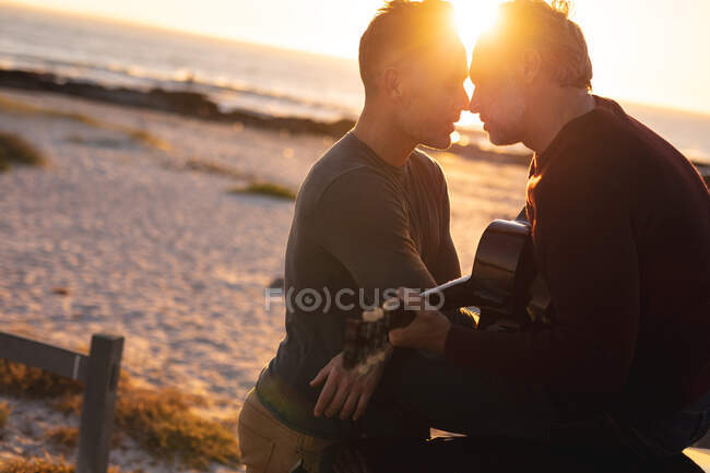 Happy caucasian gay male couple playing guitar and kissing at sundown by the sea. summer road trip and holiday in nature. — Stock Photo