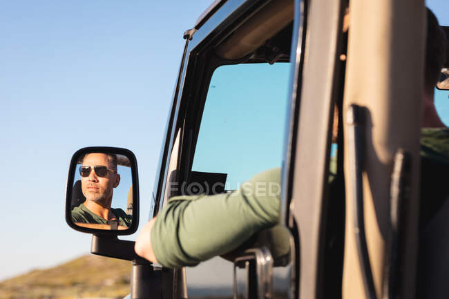Thoughtful caucasian man in car wearing sunglasses reflected in side mirror on sunny day at seaside. summer road trip and holiday in nature. — Stock Photo