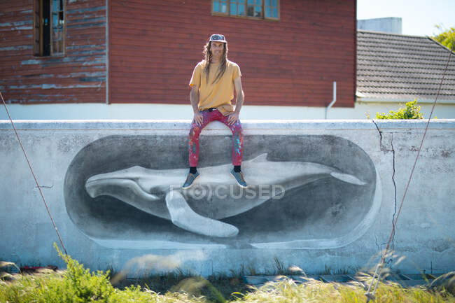 Retrato de artista masculino sentado na parede com pintura mural de baleia contra casa. arte de rua e habilidade. — Fotografia de Stock