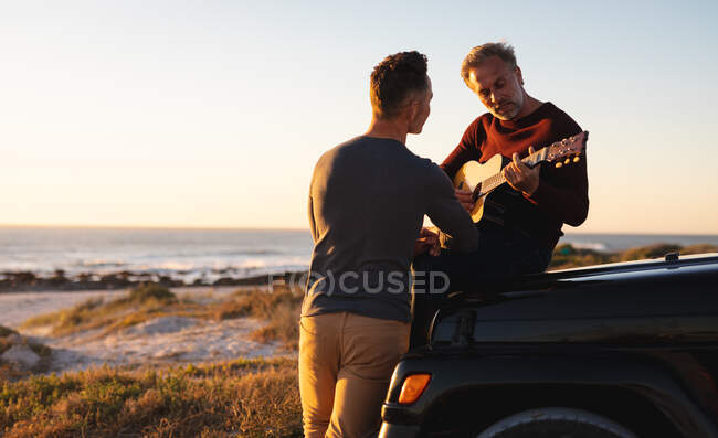 Glückliches kaukasisches schwules Paar, das bei Sonnenuntergang am Strand abhängt und auf dem Auto sitzt und Gitarre spielt. Sommer Roadtrip und Urlaub in der Natur. — Stockfoto