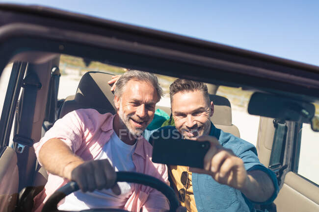 Feliz caucásico gay macho pareja tomando selfies sentado en coche en la playa. viaje por carretera de verano y vacaciones en la naturaleza. - foto de stock