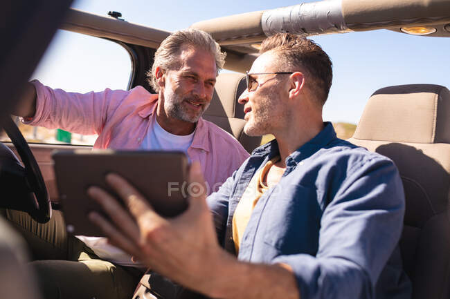 Glückliche kaukasische homosexuelle männliche Paar mit Tablette sitzen im Auto am Meer. Sommer Roadtrip und Urlaub in der Natur. — Stockfoto