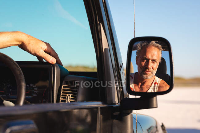 Retrato del hombre caucásico sonriente en coche reflejado en el espejo lateral en el día soleado en la orilla del mar. viaje por carretera de verano y vacaciones en la naturaleza. - foto de stock