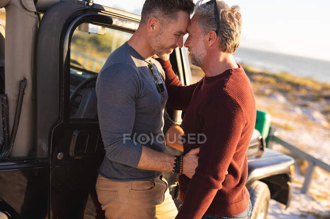 Happy caucasian gay male couple embracing by car at seaside. summer road trip and holiday in nature. — Stock Photo