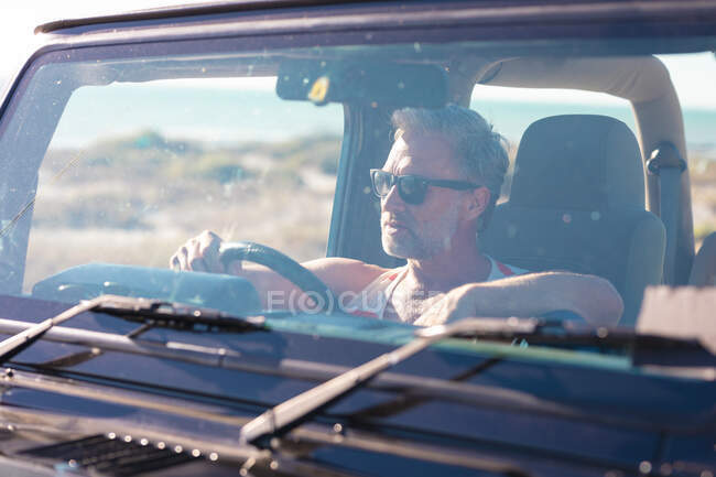 Удумливий білий чоловік у сонцезахисних окулярах сидить у машині в сонячний день на узбережжі. літня дорога поїздка і відпочинок на природі . — стокове фото