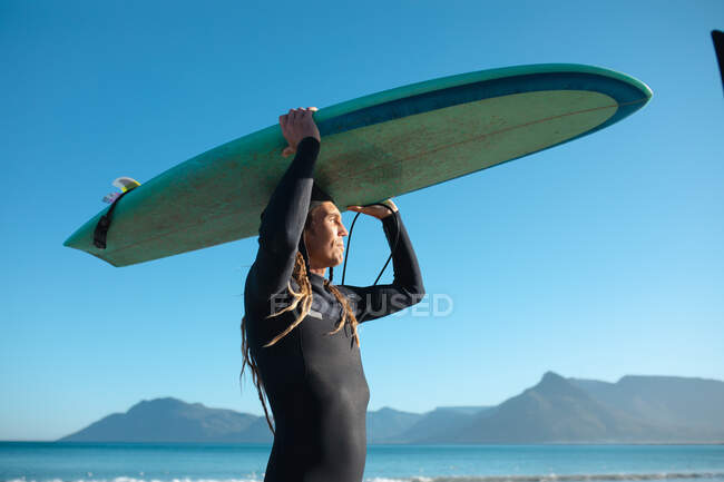 Hombre hipster llevando tabla de surf en la cabeza mientras mira hacia otro lado contra el cielo azul. pasatiempos y deportes acuáticos. - foto de stock