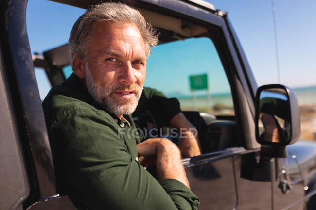 Portrait of happy caucasian man sitting in car at seaside on sunny day. summer road trip and holiday in nature. — Stock Photo