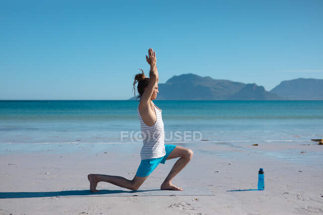 Vista laterale dell'uomo che pratica guerriero 1 posa yoga con le mani strette sopra la testa in spiaggia, copiare spazio. fitness e stile di vita sano. — Foto stock