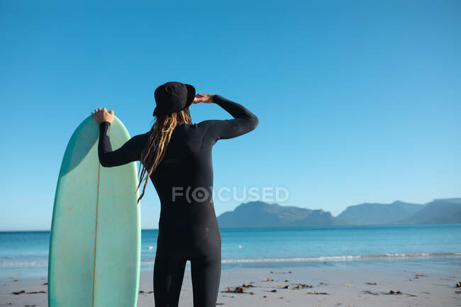 Rear view of man holding surfboard shielding eyes while looking at copy space on clear blue sky. hobbies and water sport. — Stock Photo
