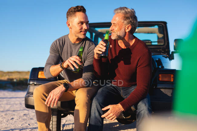 Heureux couple gay caucasien homme buvant des bouteilles de bière, assis sur la voiture au bord de la mer. road trip d'été et vacances dans la nature. — Photo de stock