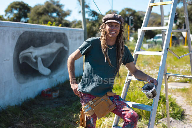 Porträt eines lächelnden männlichen Künstlers, der auf einer Leiter vor einem Wandgemälde von Walen an der Wand lehnt. Street Art und Geschicklichkeit. — Stockfoto