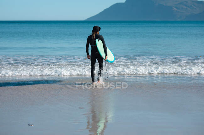 Вид сзади на человека, держащего доску для серфинга, стоя на берегу на пляже. хобби и водные виды спорта. — стоковое фото