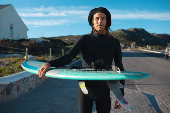 Porträt eines selbstbewussten Mannes mit Hut und Neoprenanzug, der an sonnigen Tagen Surfbretter auf der Straße trägt. Hobbys und Wassersport. — Stockfoto