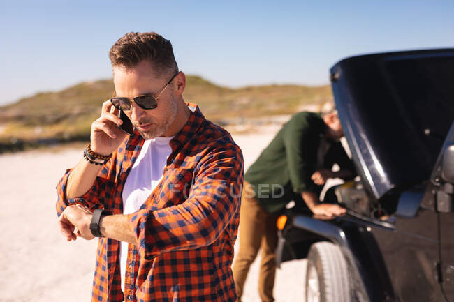 Pareja masculina gay caucásica estresada hablando en un teléfono inteligente con un coche averiado en la playa. viaje por carretera de verano y vacaciones en la naturaleza. - foto de stock