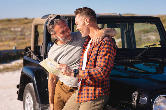Happy caucasian gay male couple embracing and reading map by car at seaside. summer road trip and holiday in nature. — Stock Photo