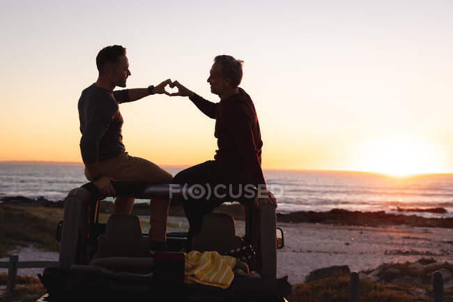 Feliz caucasiano gay masculino casal sentado no carro fazendo forma de coração com as mãos na praia ao pôr do sol. viagem de verão e férias na natureza. — Fotografia de Stock