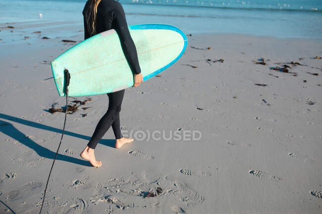 Baixa seção de surfista masculino carregando prancha na praia durante o dia ensolarado. hobbies e esporte aquático. — Fotografia de Stock