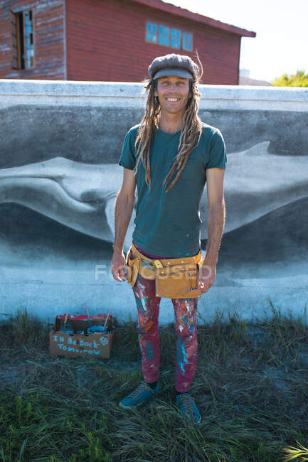Retrato sorridente de artista hipster masculino com roupas bagunçadas em pé contra a pintura mural de baleia. arte de rua e habilidade. — Fotografia de Stock