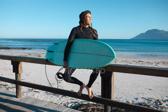 Männlicher Surfer mit Surfbrett schaut weg, während er auf einem Holzgeländer am Strand sitzt. Hobbys und Wassersport. — Stockfoto